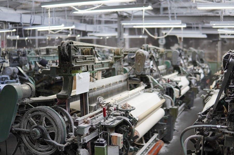 遠州特有の風合いを実現できる最大の要因は、今なお現役で動く「シャトル織機」にあります。よこ糸を巻いた舟形の部品（シャトル）が機械の中を往復し、糸の間を通りながら布を織り上げていく、最も古い形の自動織機です。空気圧などを使う最新の織機に比べ、生産スピードは20～30分の1程度。一見すると非常に非効率な機械です。しかし、糸にかかる負荷が極めて低いため、最新の織機では切れてしまうような「極めて細い糸」でも織り上げることができます。工場で動いているのは、60年前に作られた阪本式シャトル織機20台。地元の遠州製作所製で、現在は製造されていない世界的にも希少な機械です。これらを熟練の修理職人が丁寧にメンテナンスしながら、今も稼働させています。
