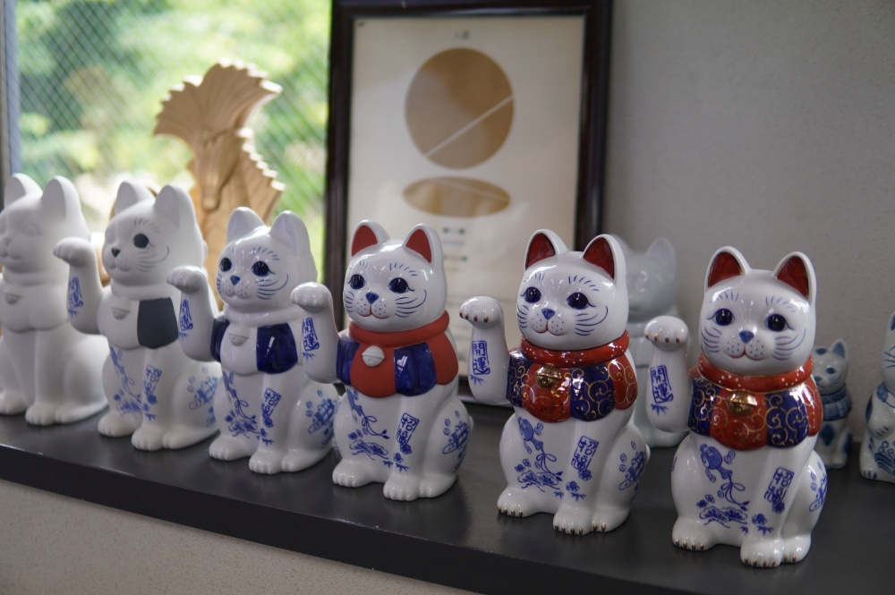 中外陶園さんの陶磁器は石膏型を用いた「鋳込み（いこみ）」という製法で生み出されています。瀬戸焼最大の特長とも言えるのが、「アタッチ」と呼ばれる技法です。明治時代にヨーロッパから伝わった型の技法が、瀬戸の粘り気のある土と非常に相性が良く、この地に根付きました。アタッチとは、本体と手などの別パーツを後からくっつける技術のことです。例えば、常滑焼の招き猫は土に粘り気がなくアタッチができないため、手が顔にくっついた丸みを帯びた形をしています。一方、瀬戸の招き猫はアタッチを用いることで手が顔に触れない独立した表現が可能となり、猫背で顔が小さい、本物の猫のような細身で美しいフォルムが特徴となっています。